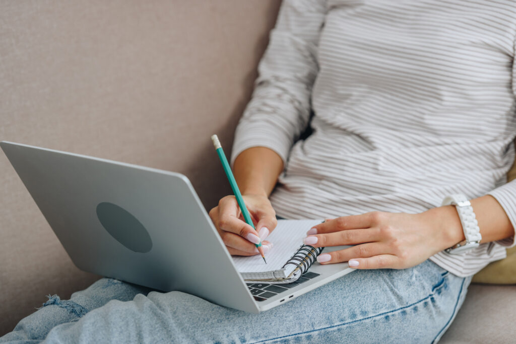 Person in a striped shirt and jeans sitting on a sofa, writing in a notebook placed on an open laptop, likely taking notes from free work from home courses.