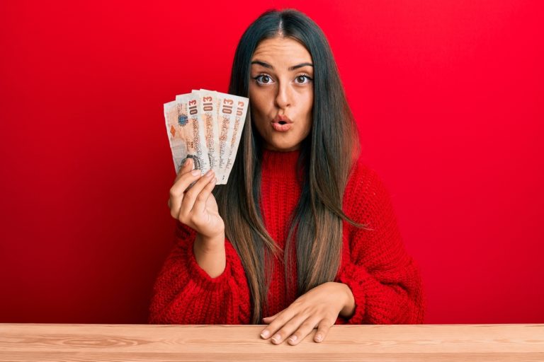 Beautiful dark haired woman holding several &pound;10 notes fanned out, looking amazed with open mouth for surprise, red background