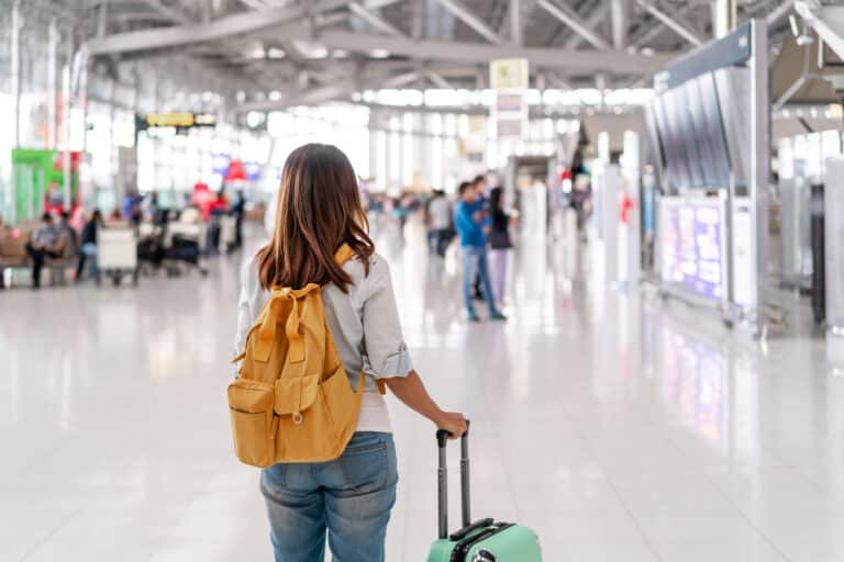 A person with a yellow backpack and a suitcase stands in an airport terminal, facing away, perhaps contemplating how one could get paid to move overseas, while other people and airport signage are visible in the background.