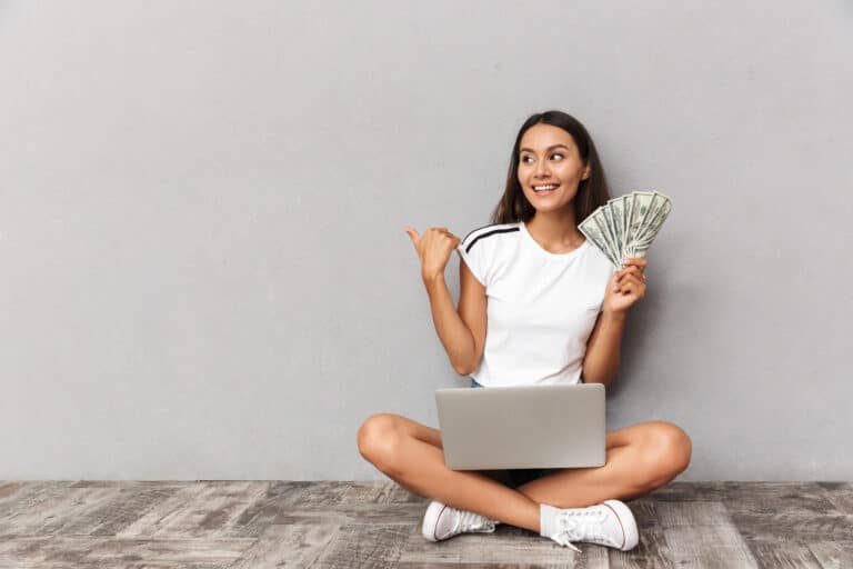 A woman sits cross-legged on the floor, holding a fan of cash in one hand and pointing with the other. With a laptop on her lap, she smiles against a plain gray wall, embodying the spirit of inspirational money quotes.