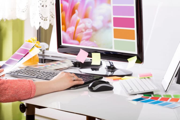Person working at a desk with a computer displaying color swatches and floral imagery, surrounded by various color samples and sticky notes, designing digital products to sell online.