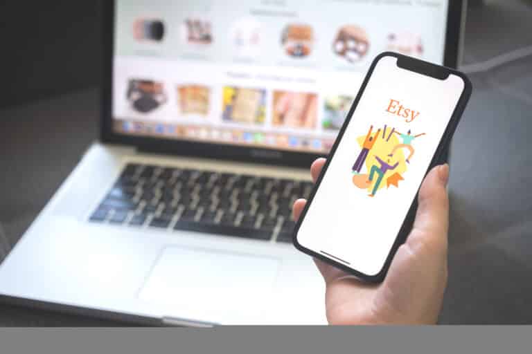 A person holds a smartphone displaying the Etsy app while a laptop in the background shows a webpage with various items, sparking clever Etsy side hustle ideas.