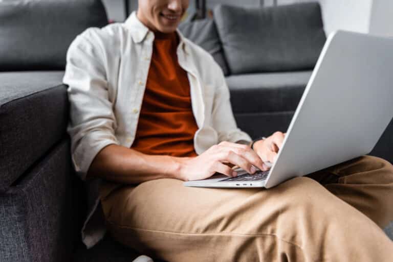 A person sits on the floor, leaning against a couch, using Outplayed Pro for matched betting on a laptop. They are wearing a white shirt over a red top and khaki pants, smiling.