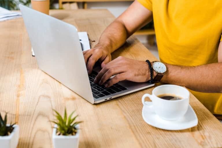 A person in a yellow shirt types on a laptop at a wooden desk, brainstorming side hustle ideas for men, with a cup of coffee and small plants nearby.
